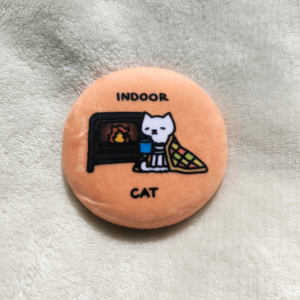 Round button pin with a cat sitting in front a fireplace illustration and text "indoor cat" on a textured surface