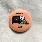 Round button pin with a cat sitting in front a fireplace illustration and text "indoor cat" on a textured surface