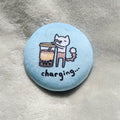 Circular button badge with a cartoon cat and bubble tea illustration on a light blue background, with text "charging...", placed on a textured surface.