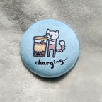 Circular button badge with a cartoon cat and bubble tea illustration on a light blue background, with text "charging...", placed on a textured surface.