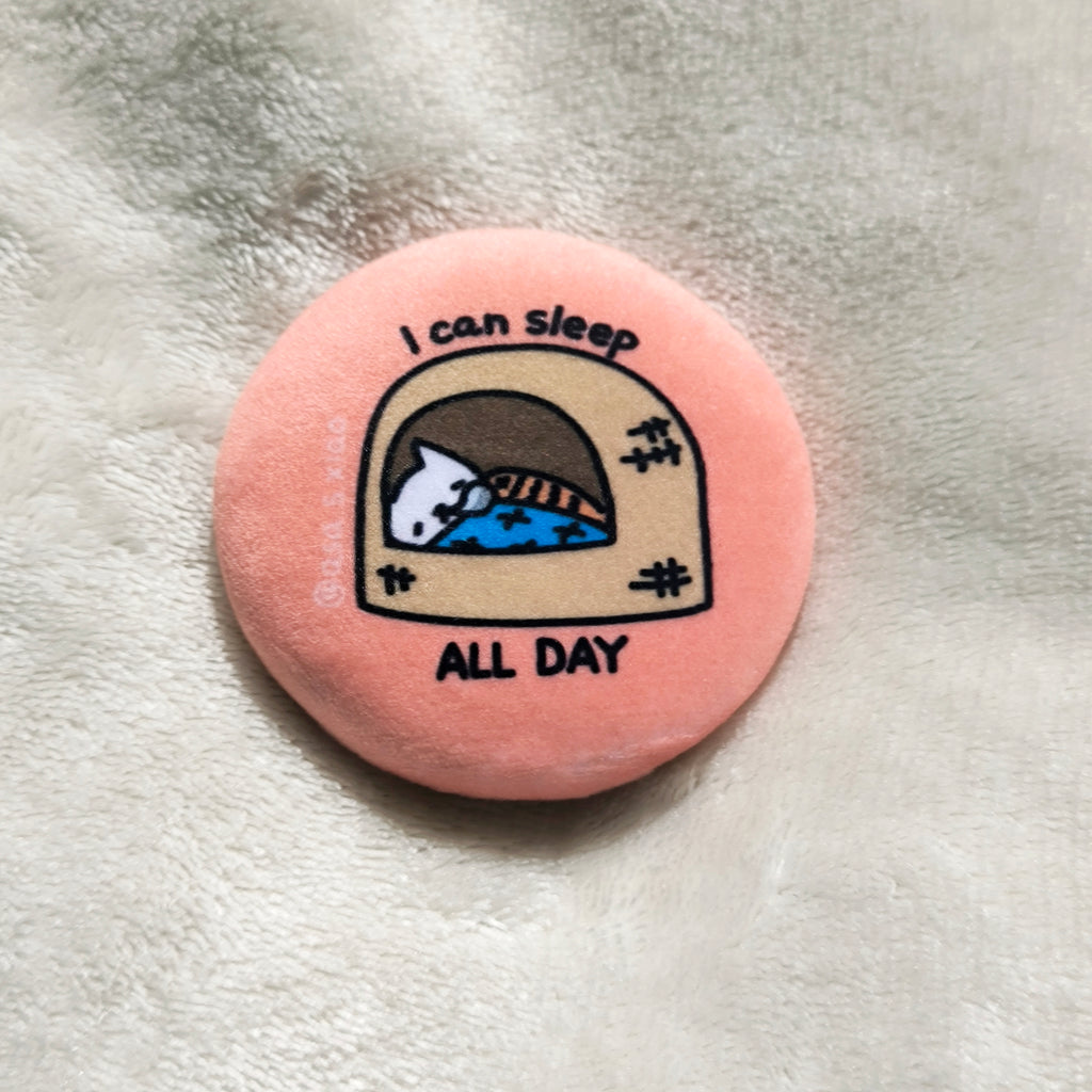 Round pink button badge with a cat illustration and text 'I can sleep all day' on a textured surface.