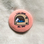 Round pink button badge with a cat illustration and text 'I can sleep all day' on a textured surface.