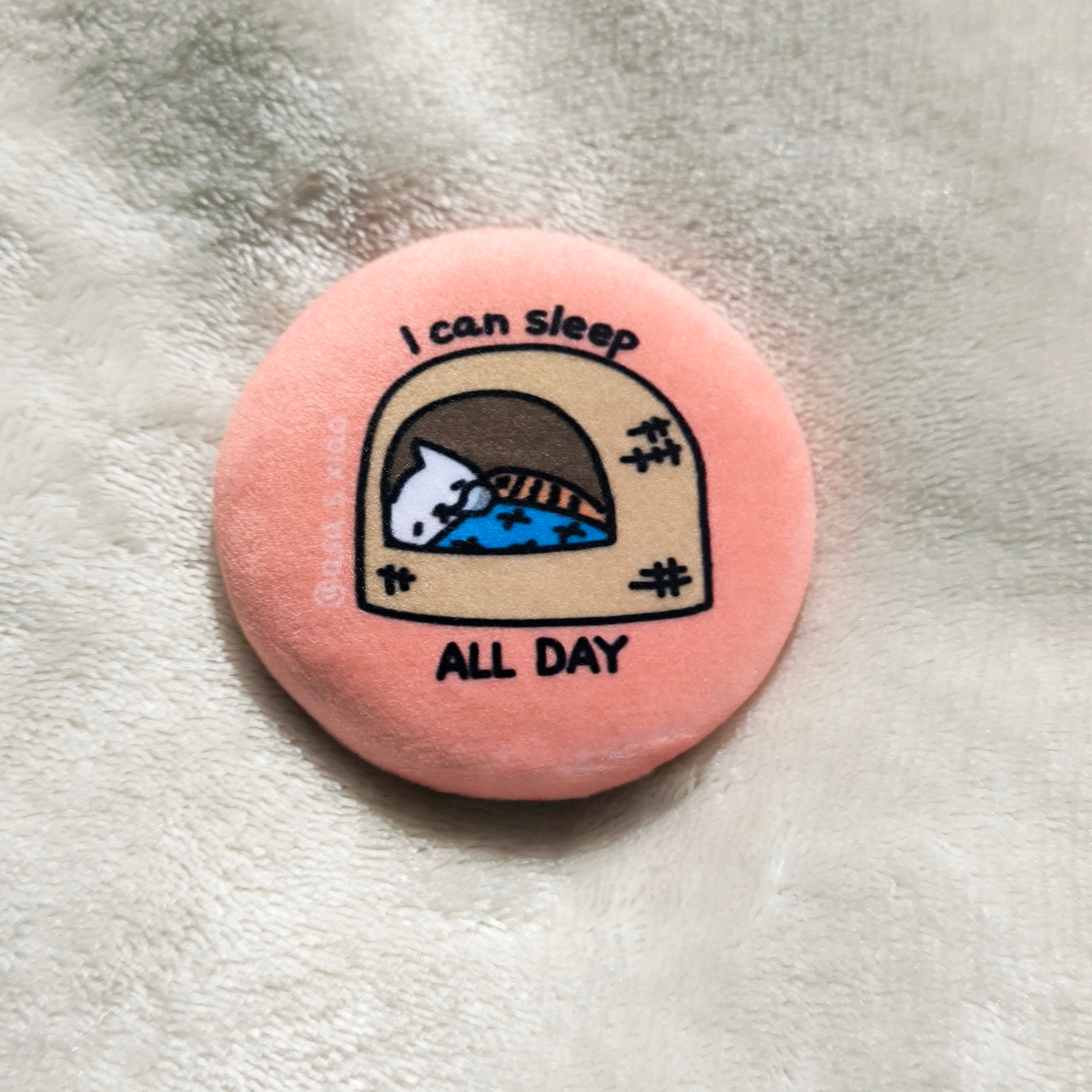 Round pink button badge with a cat illustration and text 'I can sleep all day' on a textured surface.