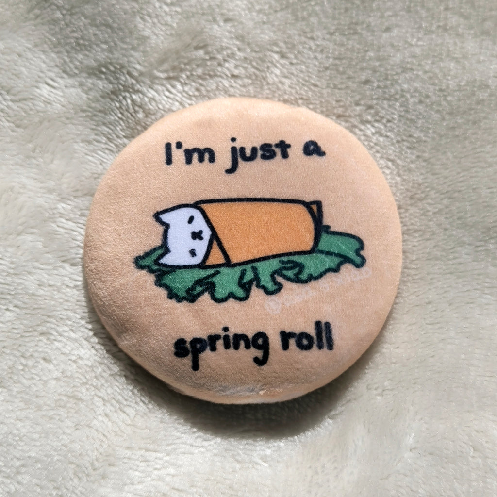 Button badge with cartoon cat wrapped in a spring roll and text 'I'm just a spring roll' on a textured surface