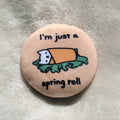 Button badge with cartoon cat wrapped in a spring roll and text 'I'm just a spring roll' on a textured surface