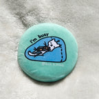 Round turquoise button with a cat illustration and 'I'm busy' text on a textured surface.