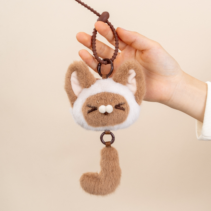 Hand holding a plush keychain shaped like a cat with a white chest on a beige background