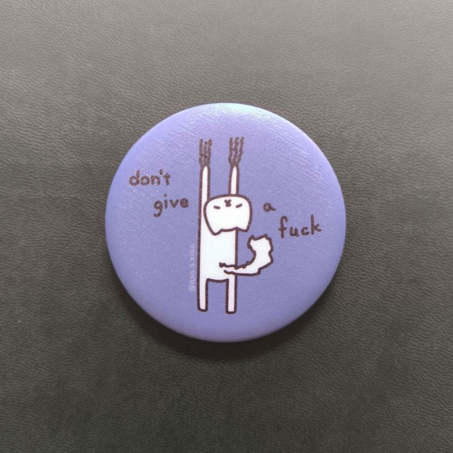 Round purple button pin with a cat character and text 'don't give a fuck' on a dark background