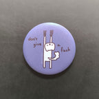 Round purple button pin with a cat character and text 'don't give a fuck' on a dark background